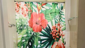 Un baño con cortina de ducha estampada con motivos de hojas y flores tropicales, un inodoro, un lavabo y una pequeña alfombra blanca en el suelo.