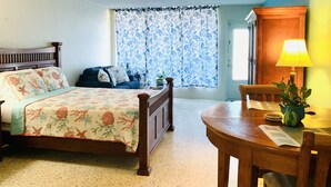 Un dormitorio con cama, mesita de noche, ventana con cortinas, una silla de madera y una mesa con lámpara.