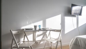 A room with a white table and two white chairs, a TV mounted on the wall, and sunlight streaming through the window.