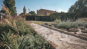 Un sentier de pierres mène à une maison de campagne rustique, avec le clocher d’une église en arrière-plan.
