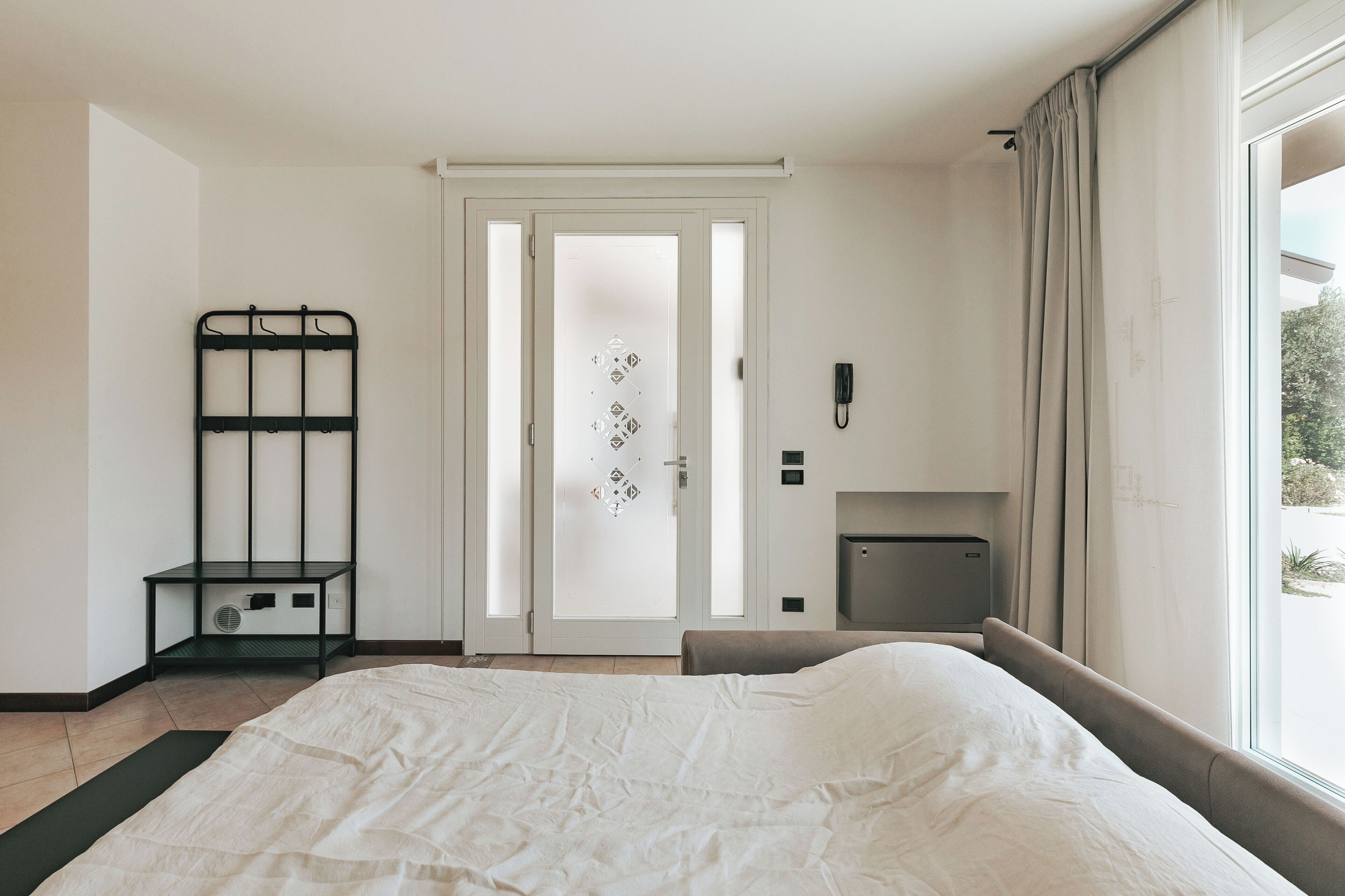 Un dormitorio con cama, mesita de noche, radiador, puerta con vidrio esmerilado y vistas por la ventana.