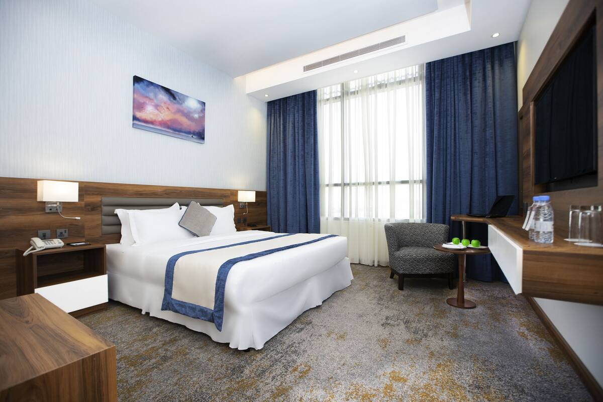 Une chambre d’hôtel avec un grand lit, un bureau avec une chaise, une petite table avec une lampe et une grande fenêtre avec des rideaux.