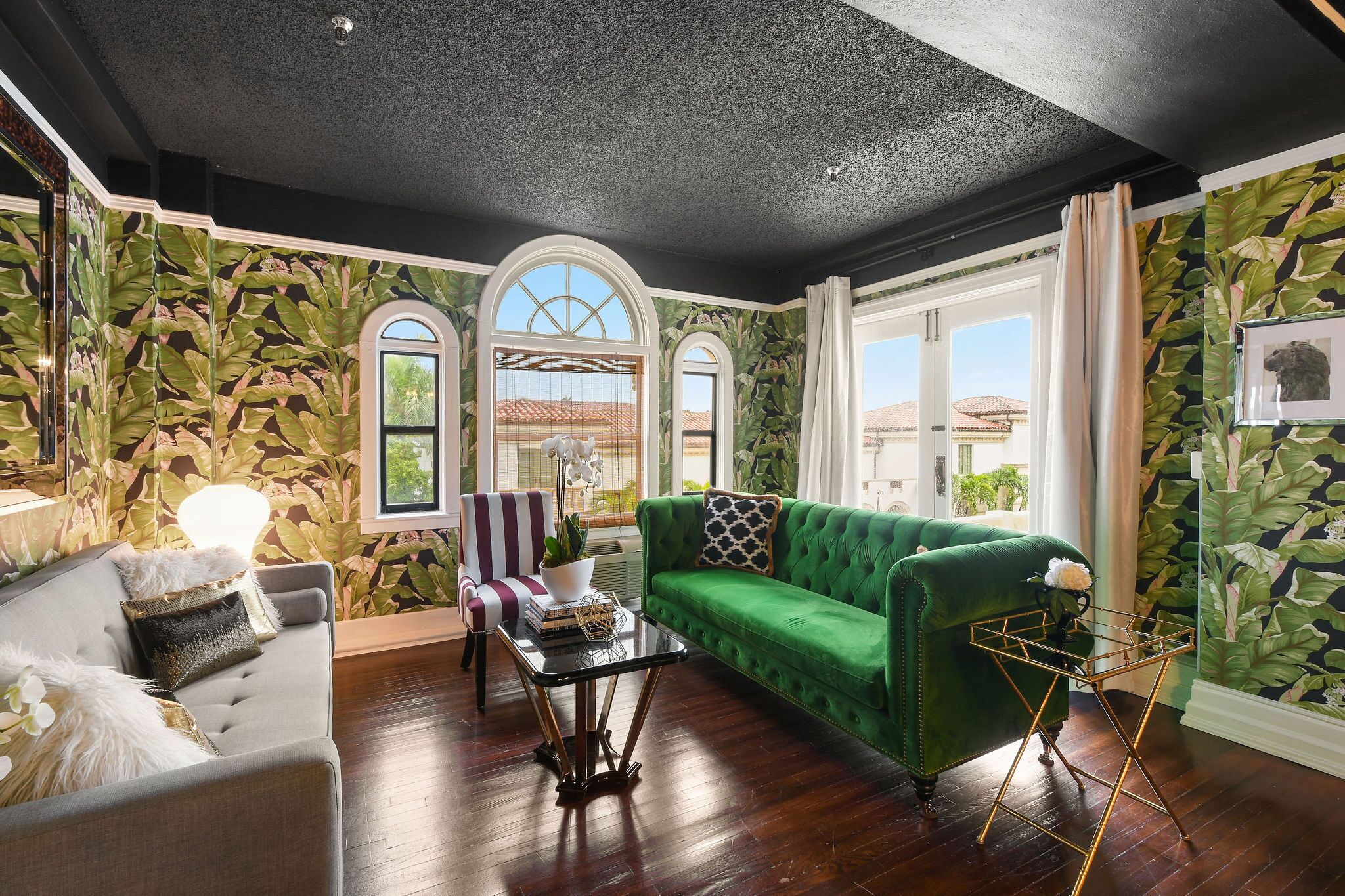 A living room with a green sofa, a striped armchair, and a glass coffee table. There are two windows with arched tops and a view of the outside.