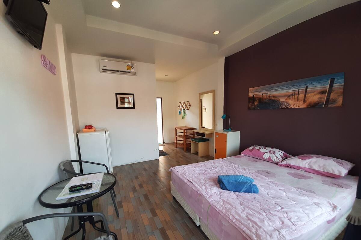 A hotel room with a bed, a desk, a chair, a small table, an air conditioner, and a painting on the wall.