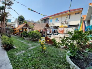 A courtyard with a thatched-roof hut, a white building with a red roof, and a variety of plants and flowers.