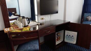 A hotel room with a wooden desk, a television mounted on the wall, a mini-fridge with water bottles, and a small cabinet with snacks.