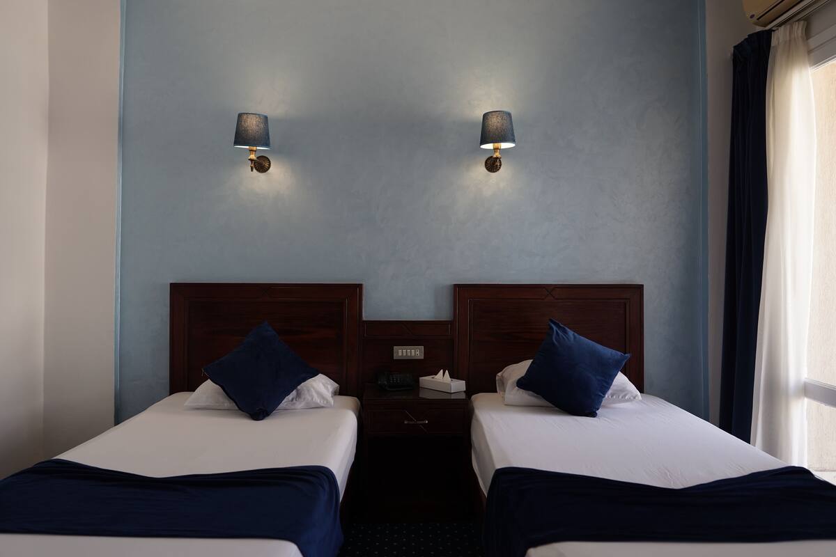 A hotel room with two single beds, a nightstand with a lamp, and a window with curtains.