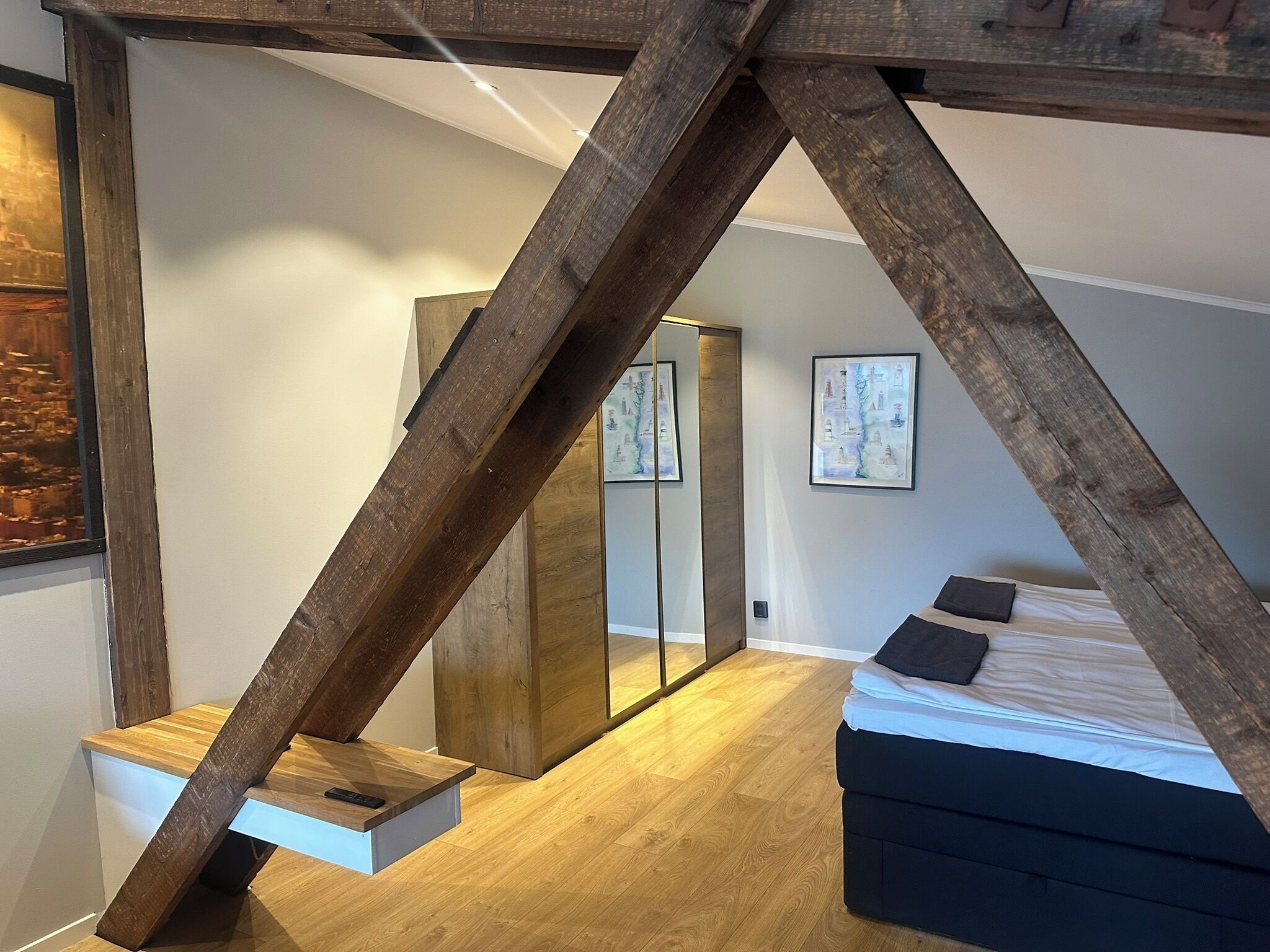A bedroom with a sloped ceiling, wooden beams, a bed, a nightstand, a mirror, and a painting on the wall.