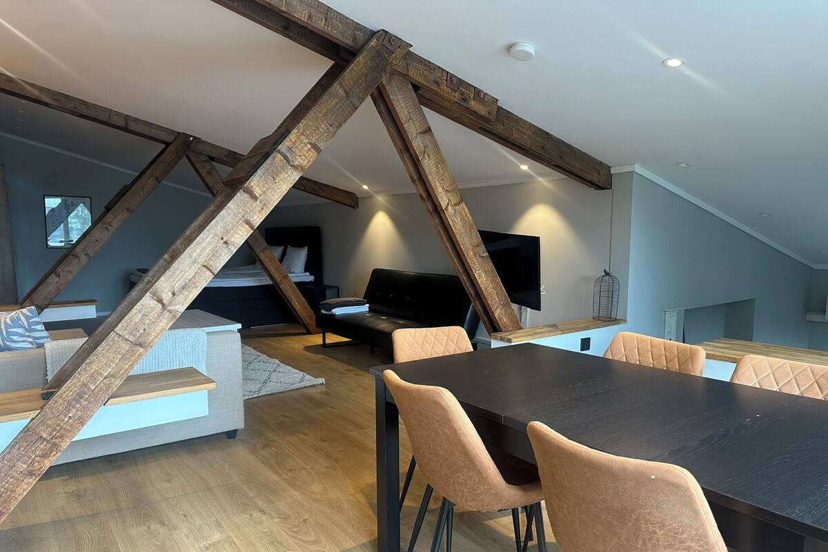 A modern loft-style room with exposed wooden beams, a dining area with a black table and cushioned chairs, a sofa, and a TV mounted on the wall.