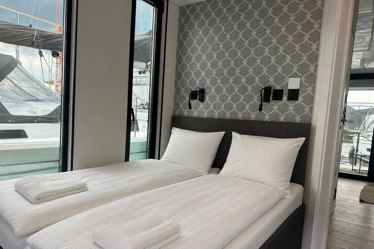 A double bed with white linens and towels, a patterned wall, and a view of boats through a window.