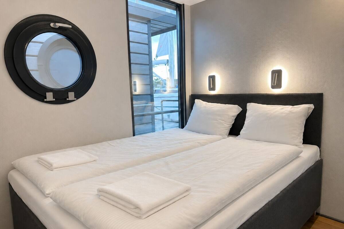 A neatly made bed with white linens and pillows, a round mirror on the wall, and a window with a view of the outside.