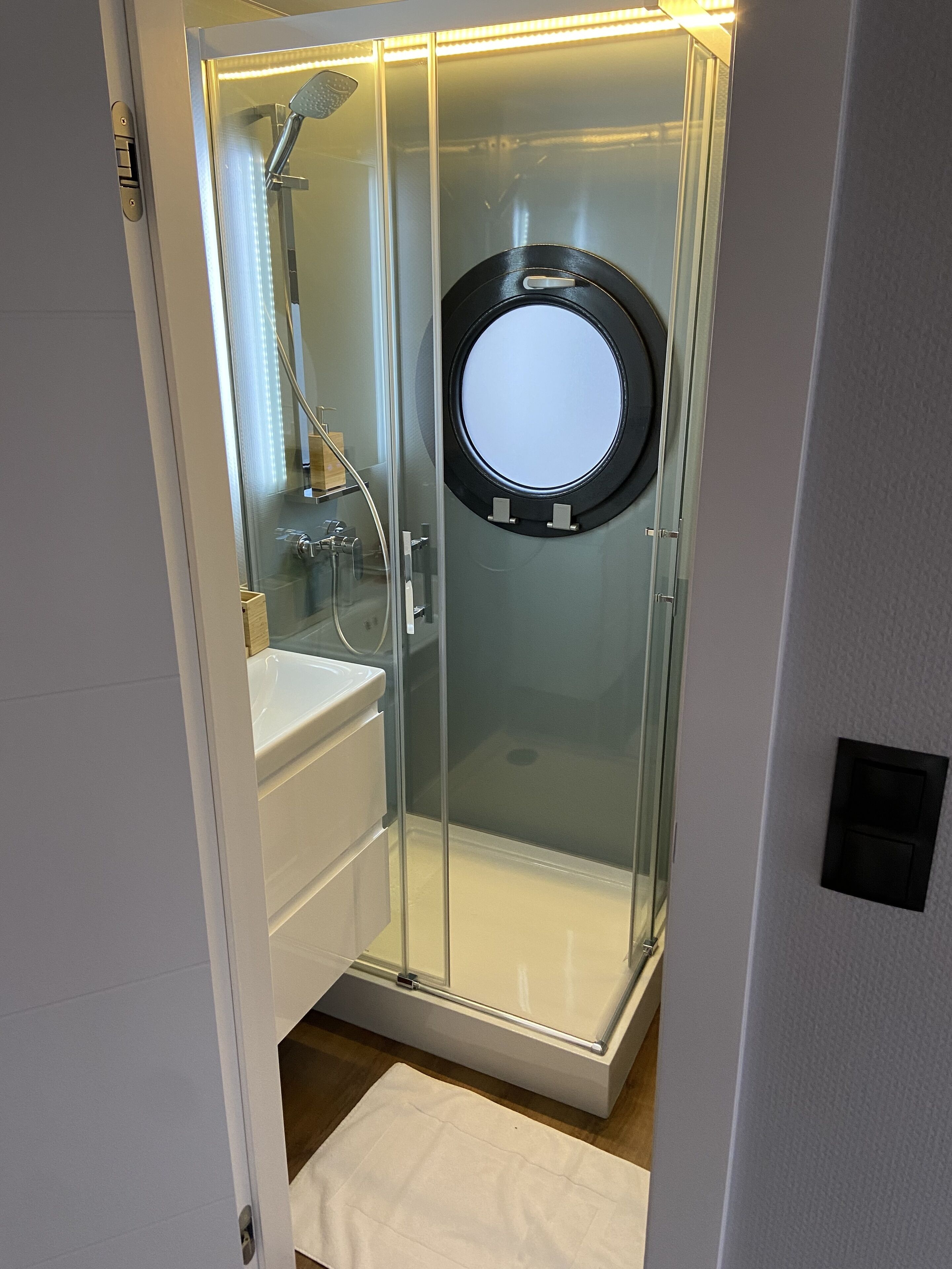 A glass-enclosed shower with a round mirror, a white sink, and a towel on the floor.