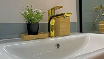 A modern bathroom with a white sink, a gold faucet, and a potted plant on a wooden tray.