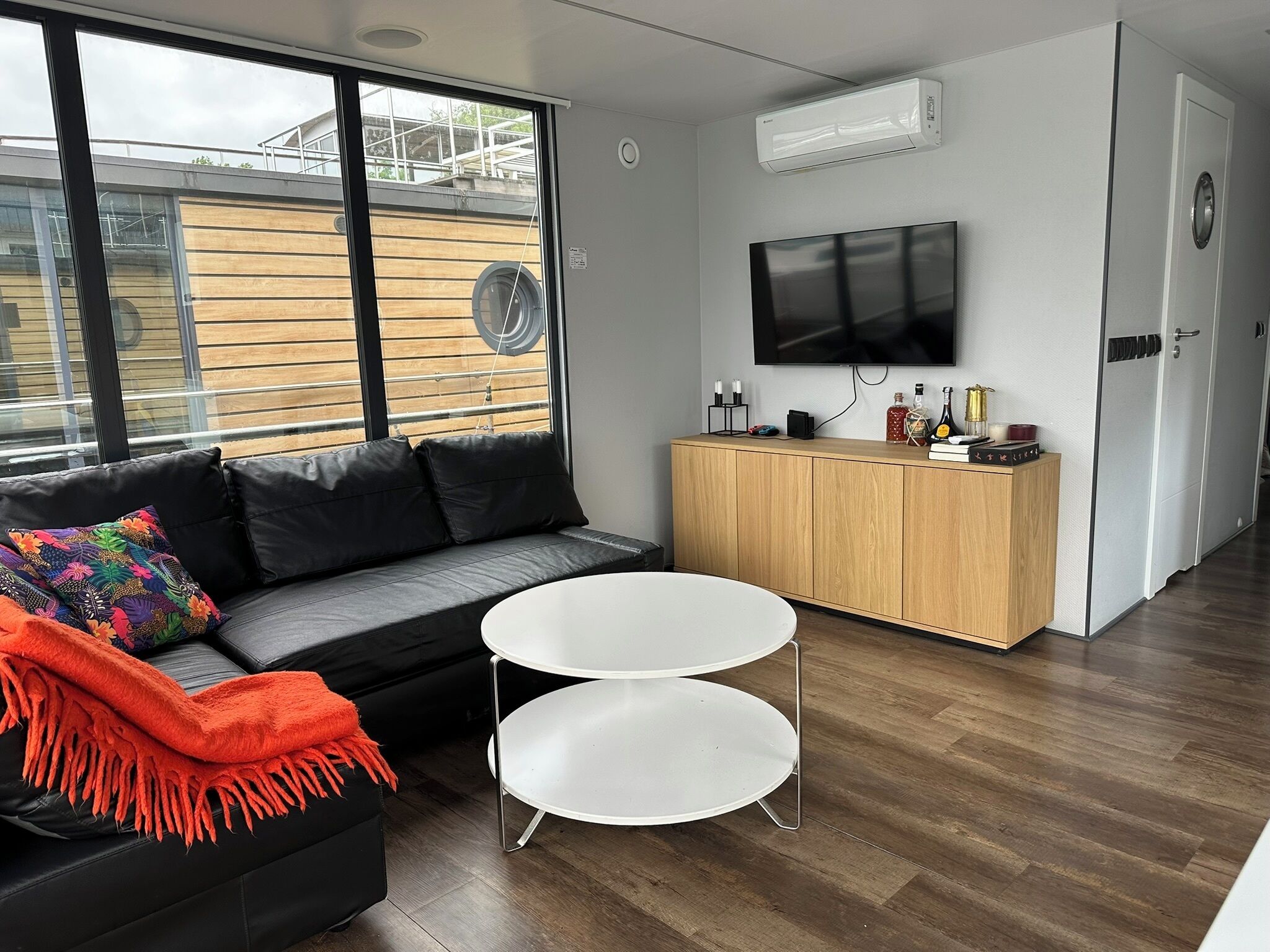 A modern living room with a black leather sofa, a white round table, a wooden cabinet, and a TV mounted on the wall.
