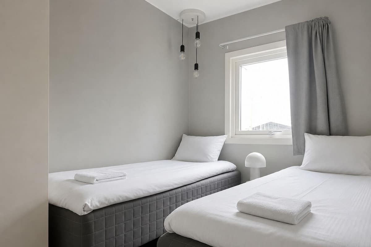 A hotel room with two beds, a window with curtains, a bedside lamp, and a view of buildings outside.