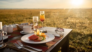 A picnic setup with a plate of food, a glass of juice, and a glass of water on a table in a field. - Cherero Camp (Serengeti)