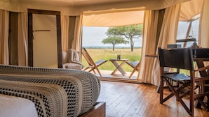 A bedroom with a large bed, a desk, and a chair, with a view of a grassy field and a tree through a window. - Cherero Camp (Serengeti)