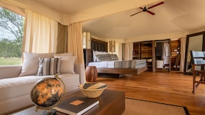 A spacious bedroom with a large bed, a bedside table with a globe and book, and a view of the outdoors. - Cherero Camp (Serengeti)