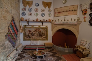 Ein traditionell eingerichteter Raum mit Steinwand, Kamin, einem gemusterten Teppich sowie verschiedenen dekorativen Tellern und Textilien.