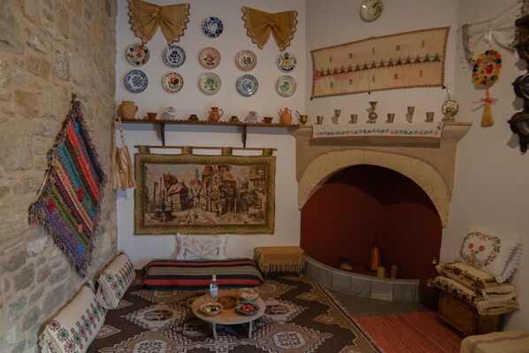 Ein traditionell eingerichteter Raum mit Steinwand, Kamin, einem gemusterten Teppich sowie verschiedenen dekorativen Tellern und Textilien.