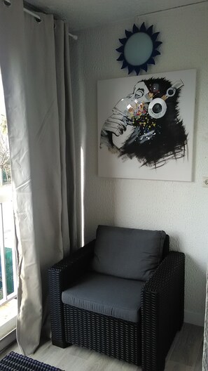 A corner room with a grey cushioned chair, a large abstract painting, a blue sun decoration, and a window with curtains.