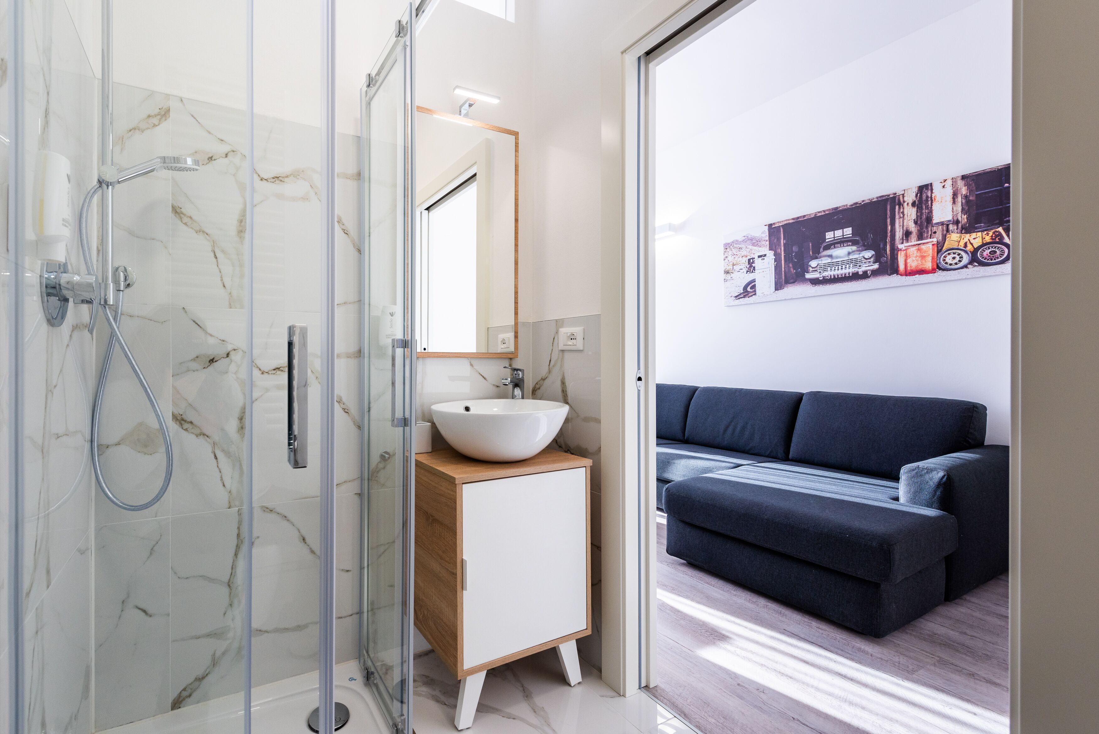 A modern bathroom with a glass shower enclosure, a white sink on a wooden vanity, and a dark-colored sofa in the adjacent living area.