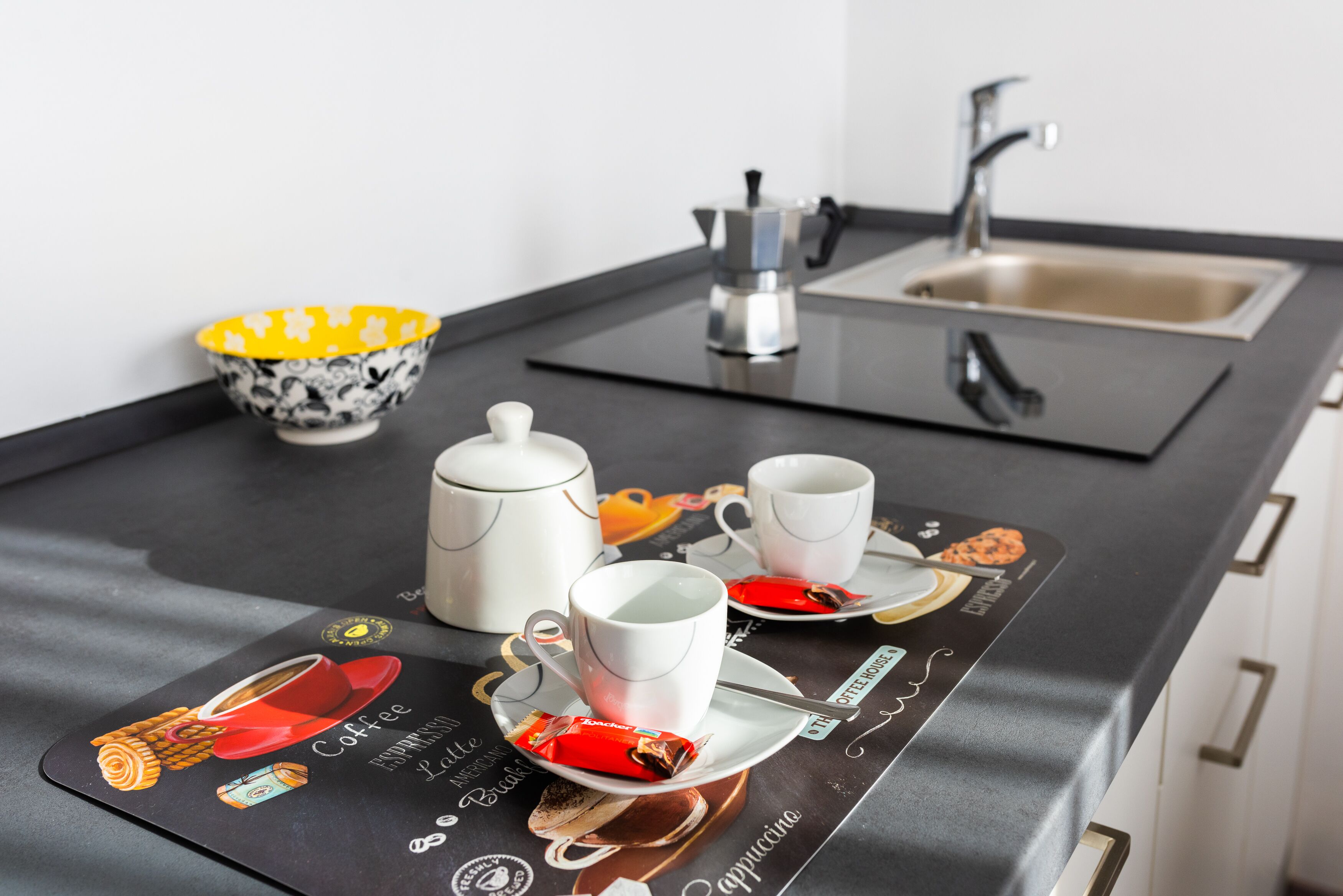 A kitchen counter with a coffee maker, two cups, a teapot, and a bowl.