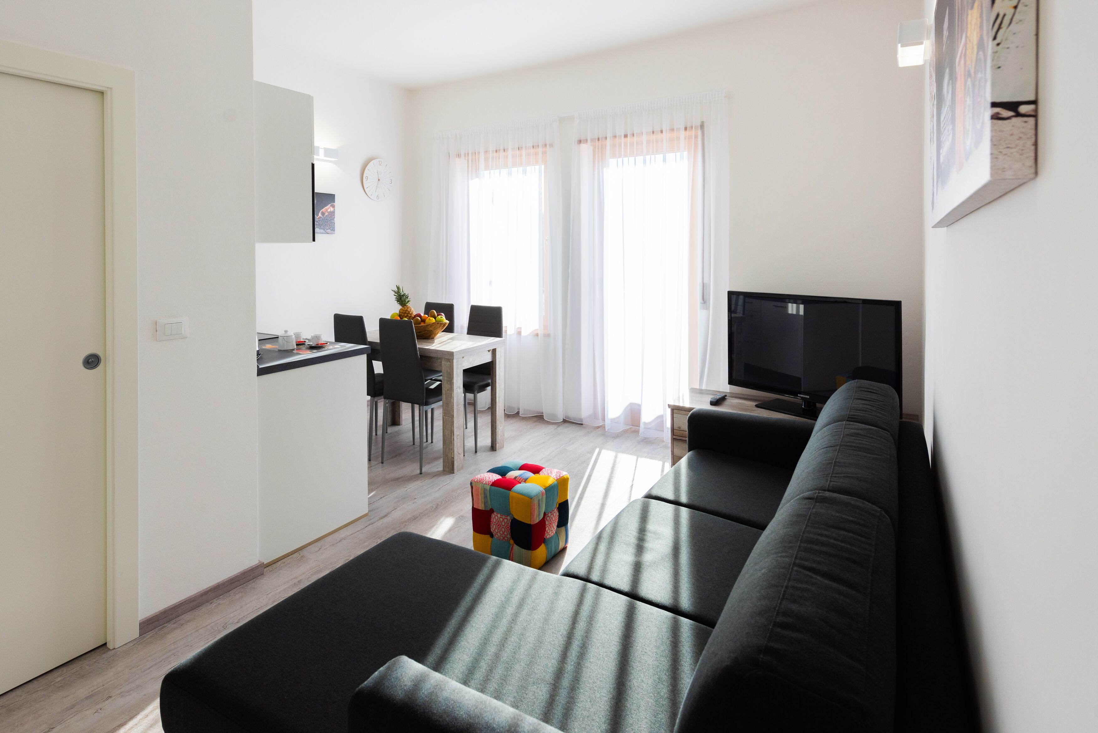 A modern living room with a black sofa, a colorful ottoman, a dining area with a table and chairs, and a television.