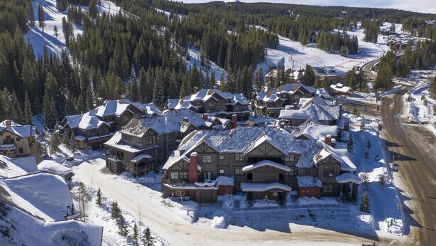 Un pueblo cubierto de nieve con casas de madera tradicionales, un camino de tierra y una pista de esquí al fondo.