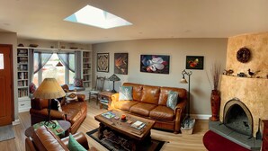 A cozy living room with a leather sofa, a coffee table, a fireplace, and a bookshelf filled with books. - Pueblo Courtyard Home on Rail Trail (Santa Fe)