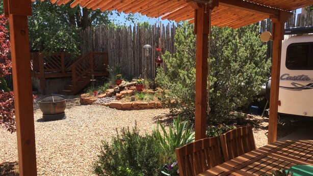 A covered patio area with a wooden table and chairs, surrounded by potted plants and a gravel ground. - Pueblo Courtyard Home on Rail Trail (Santa Fe)