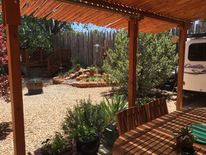 A covered patio area with a wooden table and chairs, surrounded by potted plants and a gravel ground. - Pueblo Courtyard Home on Rail Trail (Santa Fe)