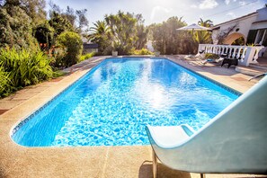 A rectangular swimming pool with clear blue water, surrounded by a tiled deck and lush greenery.