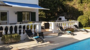 Zona junto a la piscina con sillones de descanso, una mesa y barandilla blanca.