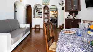 A living room with a sofa, a dining table with a patterned tablecloth, and a kitchen visible through an archway.