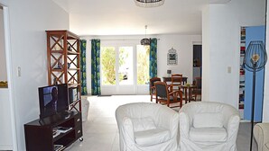Un salon comprenant un téléviseur, une bibliothèque, deux fauteuils blancs, une table à manger avec des chaises, et une vue sur l’extérieur grâce à une grande fenêtre.