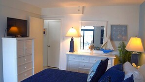 A bedroom with a TV on a dresser, a dresser with drawers, a bed with blue bedding, a dresser with a mirror, and a lamp on the dresser. - LINENS & DAILY ACTIVITIES INCLUDED! Braemar -1BR - Pool, Tennis & Fitness Center (Ocean City)