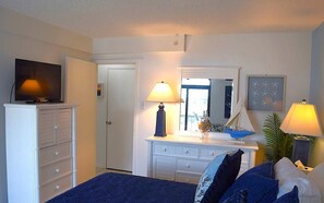 A bedroom with a TV on a dresser, a dresser with drawers, a bed with blue bedding, a dresser with a mirror, and a lamp on the dresser.