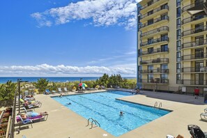 A swimming pool with lounge chairs and umbrellas, a tall building with balconies, and a view of the ocean.