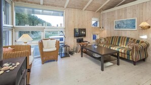 A cozy living room with a wooden ceiling, a large window, a television, a striped sofa, and a wicker chair. - Cozy home in Blokhus with WiFi (Blokhus)