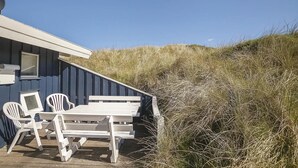 A blue building with a sloped roof, a wooden deck with a table and chairs, and tall grass in the background. - Cozy home in Blokhus with WiFi (Blokhus)
