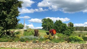 A stone wall with potted plants, a red barrel, and a grassy landscape with trees under a blue sky.