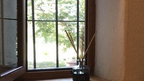 A room with a wooden floor, a window with a view of trees, and a small table with a vase and sticks.