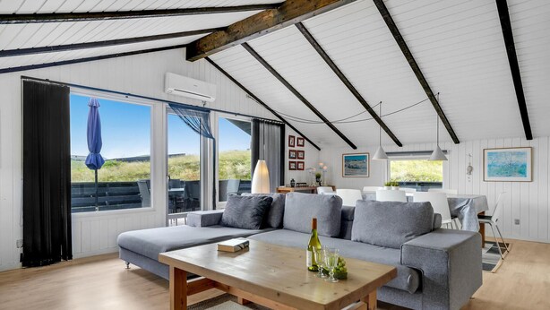 A spacious living area with a large gray sofa, a wooden coffee table, and a dining table. There are large windows offering views of the outdoors.
