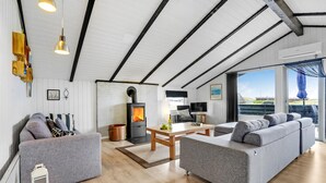 A spacious loft with a high ceiling, a wood-burning stove, grey sofas, a wooden coffee table, and a large window offering a view of the outdoors.