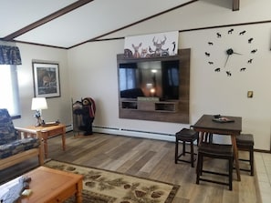 Una sala de estar con un televisor montado en la pared, un reloj, un sofá, una mesa de centro y una pequeña mesa con sillas.