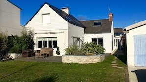 A white house with a sloped roof, a patio with outdoor furniture, and a garden area with plants and a stone planter. - Villa 6 persons, 100 m from the sea (Saint-Martin de Bréhal)