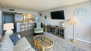 Ein Wohnzimmer mit einer Couch, einem Korbsessel, einem Fernseher und einer Küche im Hintergrund.