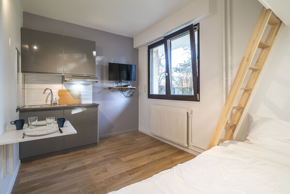 A compact room with a kitchenette, a bed, a window, and a wooden ladder.