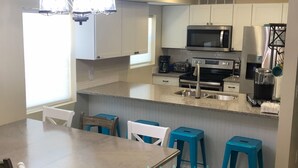A modern kitchen with white cabinets, a granite countertop, a stainless steel refrigerator, a microwave, and a coffee maker. There are two blue bar stools and a wooden dining table with white chairs. - Beautiful-condo-townhouse- Sleeps 8 - 8 Minute walk to Beach - Near Pier Park (Panama City Beach)